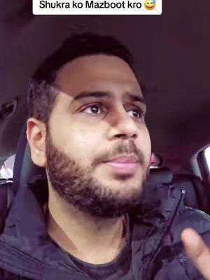 A man with a beard is sitting in a car, wearing a black jacket. The image has a slightly purple tint.