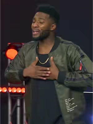 The cover image features a Black man, possibly a preacher or speaker, with his eyes closed and hands clasped over his chest. He wears a green bomber jacket, black shirt, and a gold necklace. The background is a dark stage setting with red lights.
