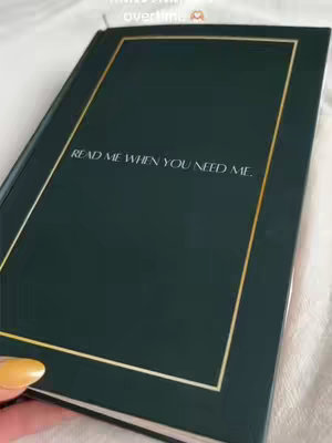 The image is a close-up of a dark green journal with a gold trim. The text "READ ME WHEN YOU NEED ME." is written in white, centered on the cover. A hand holding the journal from the bottom left, with painted yellow nails.