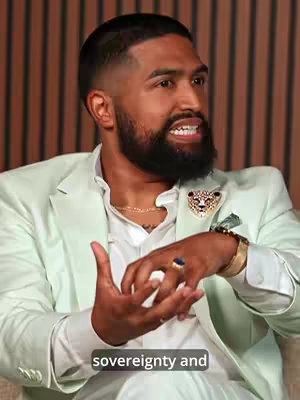 A close-up shot of a man in a light blue suit sitting down. He has a dark beard and is gesturing with his hands while speaking. A gold chain and jewel-encrusted badge can be seen on his suit. The background is a vertical striped pattern.