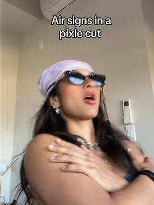 A woman with dark hair, wearing sunglasses and a headscarf, is partially visible from the chest up, with arms crossed to the side, a blurred indoor background.