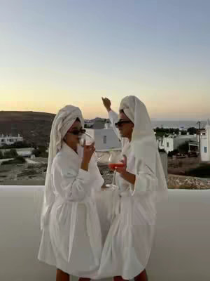 Two women in white robes and towels on their heads stand on a white balcony overlooking a coastal scene at sunset. One points upwards, while the other sips a drink. The sky gradients from soft orange to blue.