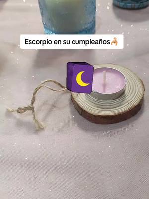 The image shows a candle with a purple candle and a wooden base. There is also a purple cube with the moon icon and string is attached to it. On the background, there is a light pink tablecloth with silver sparkles and a blue candle.