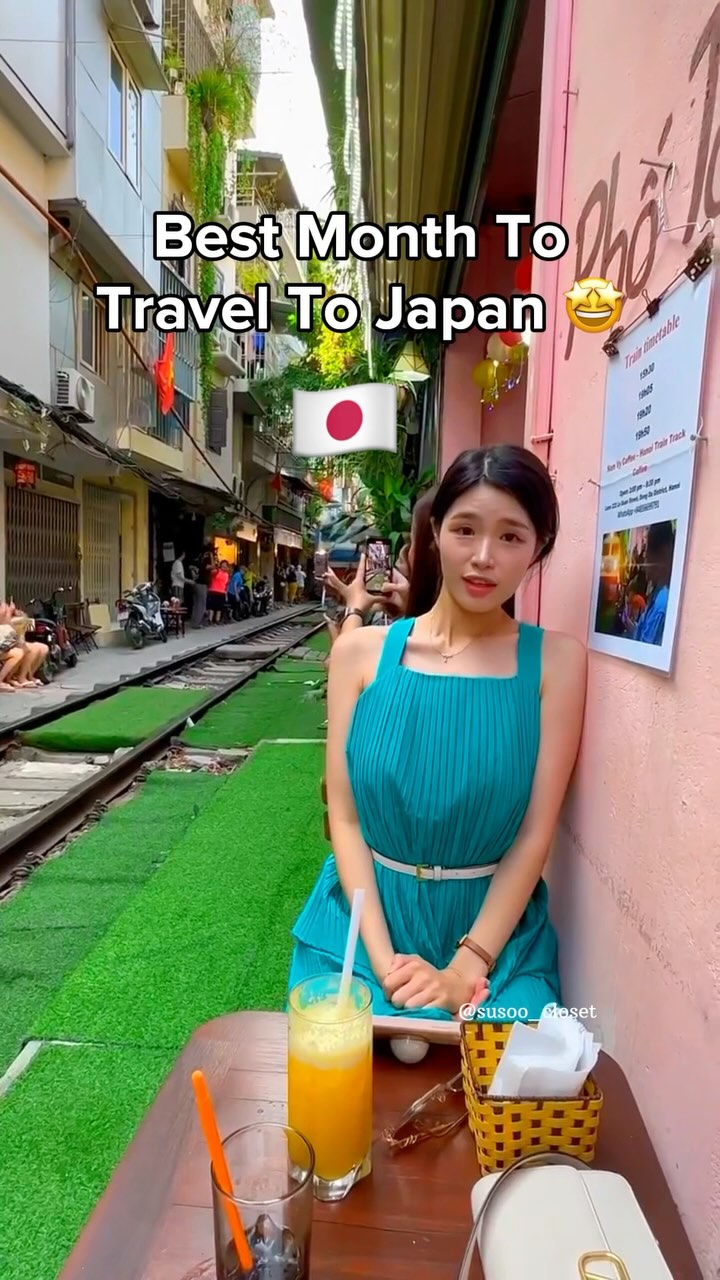 A woman in a teal dress is posing outside a cafe, in front of a pink wall. Behind her is a street with train tracks and a green surface. Text overlays the image with the text: Best Month To Travel To Japan. A drawn Japanese flag is located on the upper-right of the image.