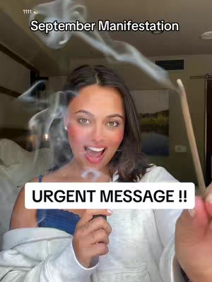 A woman with dark hair and makeup is holding a lit incense stick, with a background of blurred room details and a sign that says 'September Manifestation'. The image has a dreamy, spiritual quality with subtle smoke effects adding to the composition.