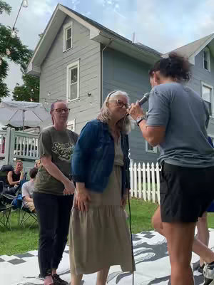 An outdoor scene featuring three women with a house and yard in the background. One woman is speaking into a microphone, with the microphone close to the face of an older woman. The third woman stands to the side. They are all standing on a patterned rug.