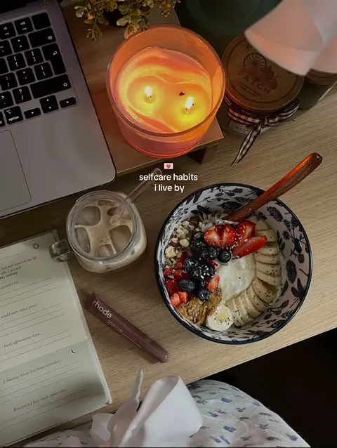 The image shows a close-up of a wooden tabletop with various self-care items casually arranged on it. A lit candle in a clear glass jar is the focal point, with two wicks casting a warm glow, suggesting relaxation. Next to it is a laptop. To the left is a glass with iced coffee and a notebook with a pen, and on the right is a bowl of oatmeal with fruit with a spoon in it. Both the arrangement and the items suggest a focus on healthy lifestyle and relaxation.