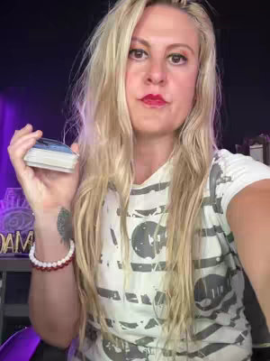 A woman holding a deck of cards, likely tarot cards, looking directly at the camera. She has long blonde hair and is wearing a white t-shirt. A beaded bracelet adorns her wrist, and a tattoo is visible on her arm. The background is dark, with a purple glow and some obscured objects, which could be decorative elements or furniture.