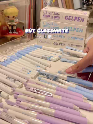 The image shows a desk or table filled with various gel pens. The pens are arranged in a visually appealing way to showcase the stationery. In the background, there are boxes of gel pens stacked on top of each other. A small figurine of a character is visible. The scene uses soft lighting and is likely setup to showcase the product.