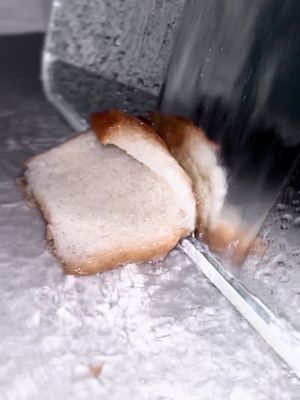 The image shows a close-up of a slice of bread being cut. The bread slice is light colored with a slightly browned crust. A shiny knife angles in, cutting the bread. The background is blurred and appears to be a metallic surface, with abstract reflections playing.