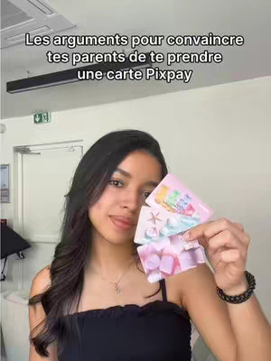A young woman with long, dark hair is holding up three colorful cards in front of her, smiling at the camera. She's wearing a black top, and the background is a neutral interior space.
