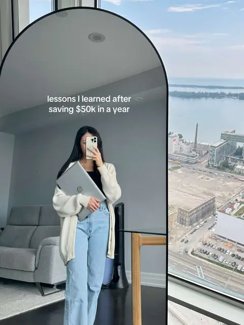 A person is reflected in an arched mirror, holding a silver laptop, in front of a window with a city view. The person wears a cream-colored cardigan, a black top, and light blue jeans and is holding their phone up to take the picture.