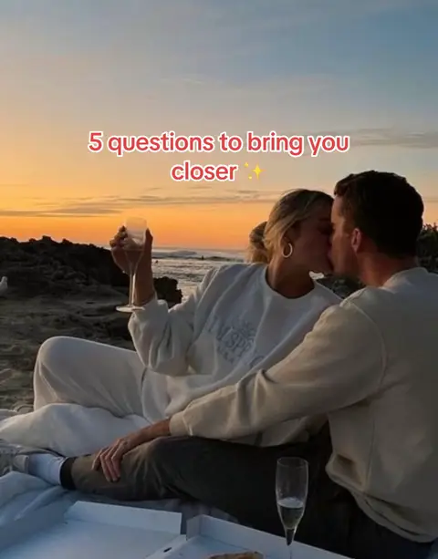 A couple kissing on a beach at sunset, with text overlayed on top. The sky is filled with orange and yellow hues. A woman holds a glass of champagne. The foreground includes rocks and a picnic setting.
