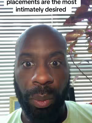 A close-up of a Black man with a shaved head and a full, dark beard. He has a slightly surprised expression, with wide eyes looking directly at the viewer. Horizontal blinds are visible in the background.
