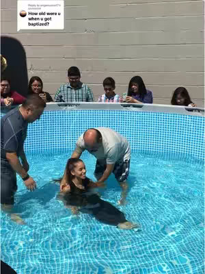 The image shows a baptism ceremony in a backyard pool. A young woman in a black outfit stands in the blue water, supported by two men, one holding her as she smiles. Around the pool's edge, several people watch. Text in the top left corner shows a question: "How old were u when u got baptized?"