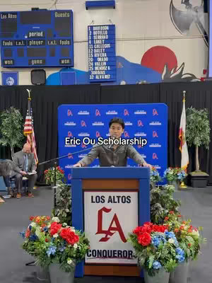 A man stands behind a podium on a stage, addressing an audience. The podium has the words "Los Altos" and the school's logo on it. There are floral arrangements in front of the podium. In the background, there are signs for a scoreboard, and other decorations. Overlayed on the image is the text "Eric Ou Scholarship". The color palette is dominated by blue, and red.