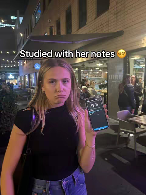 A woman stands in front of a building with a sad expression, holding a phone displaying a percentage. The background shows a street scene with some people.