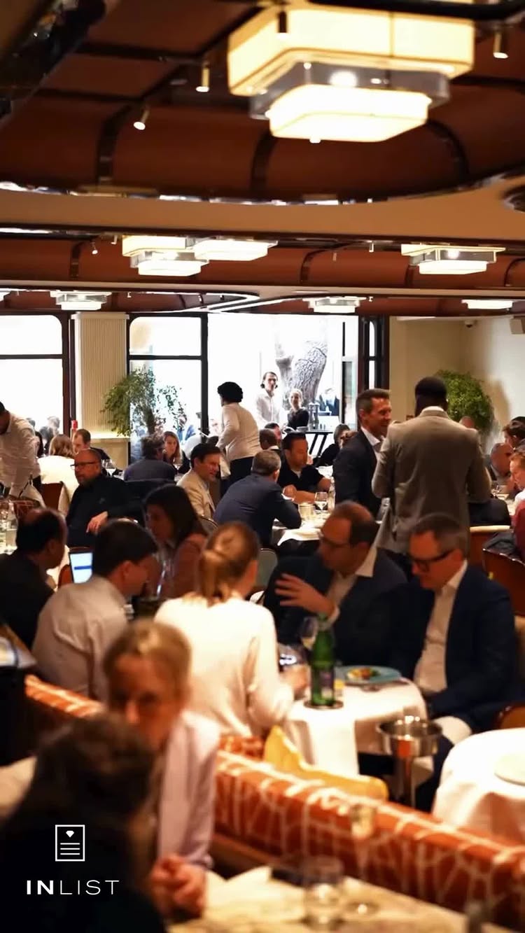 The image shows a bustling restaurant interior. Tables are set with white tablecloths, occupied by people dining and conversing. The lighting is warm, with multiple overhead fixtures illuminating the scene. The perspective is from a slightly elevated angle, capturing the full scope of the dining area. In the bottom corner, the InList logo is visible.