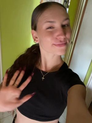 A selfie-style image showing a young woman in a black crop top in a well-lit indoor space. The woman is positioned near the edge of the frame with her arm forward, waving at the camera. She has long, dark hair pulled back from her face. The background is a light green wall and part of a door frame.