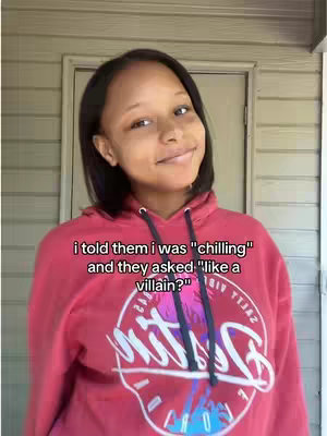A young woman with braided hair smiles at the camera, wearing a red hoodie with a graphic design. The background shows a door and siding.