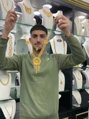 A man stands in front of a glass display filled with gold jewelry, holding up a large gold necklace. The man is facing the camera, wearing a green long sleeve shirt. The lighting is bright, highlighting the gold jewelry.