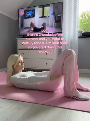 A woman is lying on a pink yoga mat in a room, seemingly indoors. She is wearing a white long-sleeved shirt and pink pants, with her legs raised in a pilates pose. Behind her is a white dresser with a TV on top, which displays a pilates workout. Text overlayed on the image says 'pov: there's 2 weeks before summer and you want to quickly tone & slim your body so you start doing this workout.'