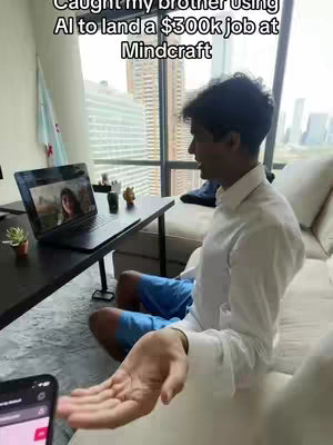 A man sits in a modern apartment setting, appearing to be on a video call. He is seated on a beige couch with a small black table in front of him holding a laptop displaying a video call. City buildings are visible through a large window in the background.