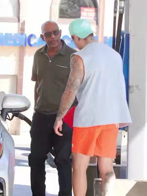 Two men are standing near a gas pump at a gas station. One man, wearing a green cap and orange shorts, is talking to another man who is wearing a polo shirt and sunglasses.