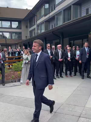 A group of people, mostly men in suits, are walking near a building. The central figure, a man in a dark suit, is walking towards the viewer with a slight smile, surrounded by others in formal attire. The background includes a building with large windows and a cloudy sky.