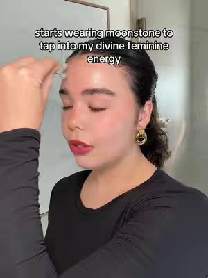 A selfie-style shot featuring a young woman with fair skin. She's positioned off-center, looking slightly upward with a contemplative expression. She wears reddish lipstick and small gold earrings. Her hair is pulled back, and she wears a black long-sleeved top.

The background is a simple interior space with a cabinet visible to the right.