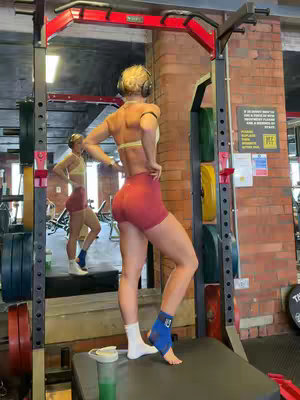 A woman stands in a gym, facing a mirror, with her back to the camera. She wears a red workout shorts, a white sports bra. She has a leg brace on her ankle, and a headphone on her head. The lighting in the gym is bright, focusing on the woman. In the background, a weight rack and exercise equipment can be seen.
