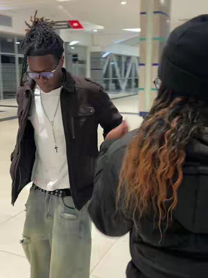 The cover image shows a man in a dark jacket and sunglasses, facing slightly downward while talking to someone off-camera. The clothing is dark, modern and stylish. The scene occurs in what looks like a mall or large indoor space. Another person with long hair, mostly out of focus, fills the right side of the frame.