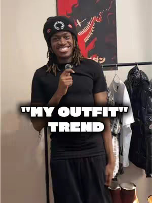 A smiling Black man wearing a black beret and black shirt stands in front of a rack of clothes. White text overlays the image, reading, "MY OUTFIT TREND," in a bold, sans-serif font. The man has dreadlocks and is holding a small microphone. The background includes a poster and a clothes rack.