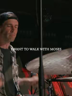 A shot from a live music performance. A man in a black cap and plaid shirt is playing drums. Text overlayed on the image reads, 'I WANT TO WALK WITH MOSES'. The background is dark with red lighting.