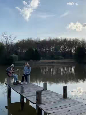 A serene outdoor scene depicts a wooden dock extending over a calm body of water, likely a lake or pond. Two people are seated on the dock: a person on the left is playing a guitar, while another is seated nearby, seemingly listening or observing. The sky is partly cloudy, with soft sunlight filtering through. The overall composition evokes a sense of peacefulness and contemplation.