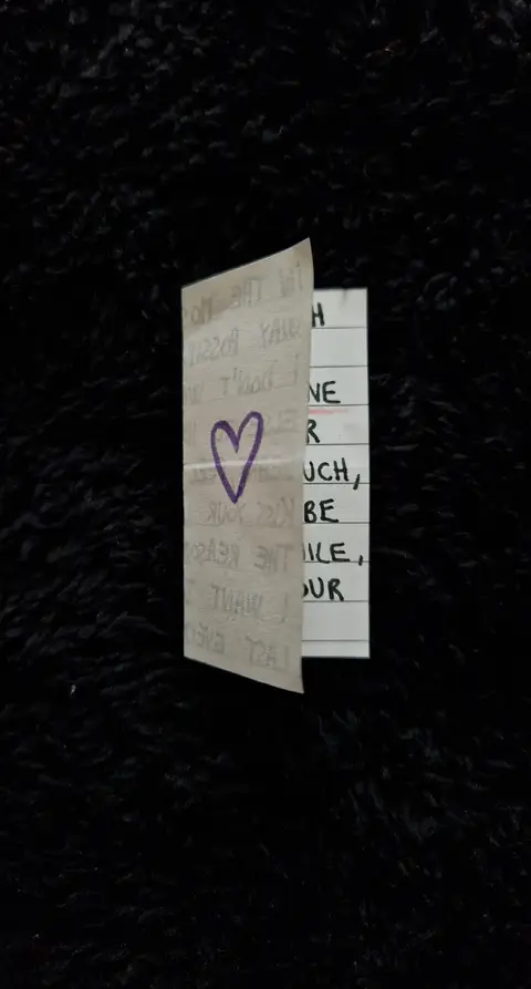 A vertically-oriented image featuring a piece of folded paper against a dark, textured background that appears to be black fabric or a similar material. The paper is off-white, and its edges are slightly worn, suggesting it has been handled.
