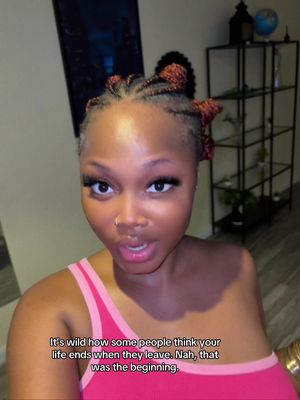 A selfie-style shot featuring a young woman with dark hair styled in braids and knots. She's wearing a deep-pink tank top. The background is a room with neutral-toned walls and a dark piece of furniture. Text overlayed at the bottom reads, "It's wild how some people think your life ends when they leave. Nah, that was the beginning."