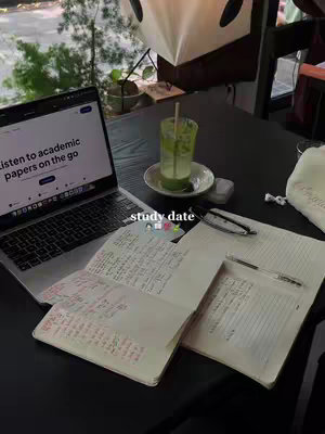 A photograph of a workspace, likely a cafe, with a table and various items related to studying. A laptop displays text, next to a notebook with handwritten notes. A glass with a green drink sits in the center, and beside the laptop, the text 'study date' appears.