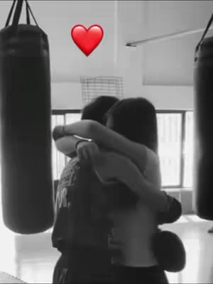 A black and white photo shows two people embracing in a gym-like setting. A red heart emoji floats above them. The scene is dark with some light coming from a window. Punching bags are visible.