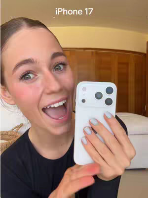 A woman with a surprised, excited expression holds a white iPhone 17. The top of the image reads "iPhone 17". The woman has rosy cheeks and light blue nail polish. The background suggests an interior setting with a bed.