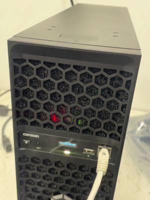 A close-up photo of a black computer server case with a mesh front panel featuring a honeycomb pattern. The case has the "canaan" logo.  There are lights visible behind the mesh and a white ethernet cable plugged in on the lower right.  The background is slightly blurred.