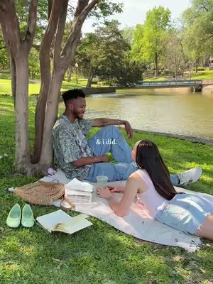 A couple is sitting on a white blanket under a tree in a park beside a lake. The man sits with his back against the tree trunk, and the woman is lying across the blanket, facing him. There are items on the blanket such as a book, a straw bag, and a container, and the couple is smiling and looking at each other. Trees, grass, and the lake are visible behind them.