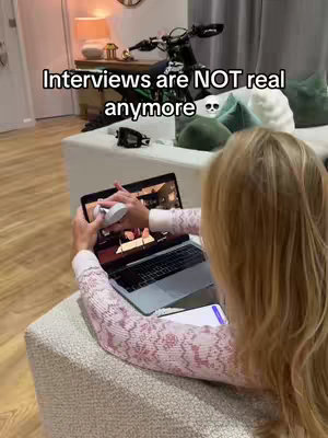 A woman is holding a laptop and using headphones while sitting on a couch. The frame shows a room interior with a sofa, a motorcycle in the background over the sofa, and additional decorative items.
