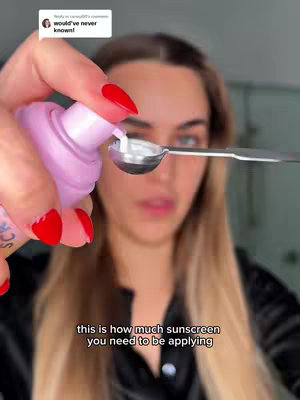 A selfie-style video cover image shows a woman holding a pink sunscreen bottle and dispensing the product onto a measuring spoon. She has long blonde hair and is looking into the camera. The background is blurred. Text at the bottom says 'this is how much sunscreen you need to be applying.' There is a comment overlay from a viewer.