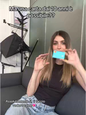 A woman in a black top and jeans holds a credit/debit card, smiling at the camera. Text overlays on the image ask if a card is possible for those 10 and up and responds saying they will show how. The background is a room with lighting equipment, a couch, and a door.
