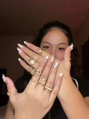 The cover image shows a young woman with long nails showing off various gold rings on both hands. The nails are long, pointed, and have a white tip French manicure with flower decorations. Her hands are in the foreground, and the background is blurred, suggesting a dimly lit indoor setting. She is looking at the camera through an opening in her hands.