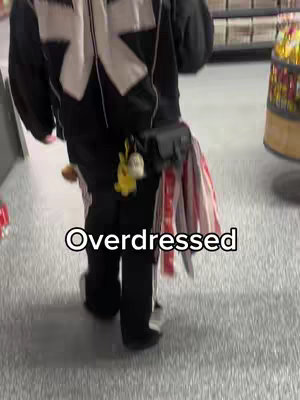 A person is walking in what appears to be a store. They are wearing black pants, black and white jacket, and a fanny pack that has several keychains hanging off of it. The person is also carrying a striped bag. The floor is a light grey color with the text 'Overdressed' written in white at the bottom of the image.