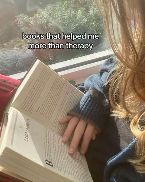 The image shows a person holding a book open, with the pages of the book visible, sitting near a window with greenery visible outside. The person is partially visible, their face obscured by their hair.
