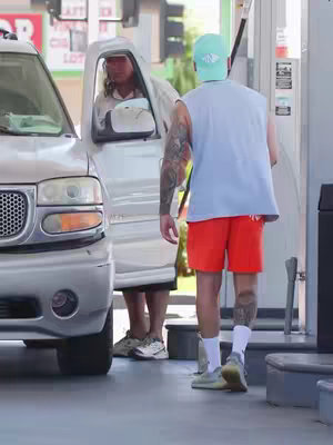 The cover image features a man standing near a silver SUV at a gas station, seen from the back. Another man, viewed from the waist up, chats with the driver of the SUV, whose door is open. The man in the forefront  wears casual attire: a light blue sleeveless top, red shorts, and a light green cap. One arm has a full sleeve tattoo. The setting is outdoors on a sunny day. The overall composition portrays a slice of life scene.
