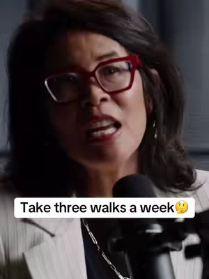 A woman with dark hair and red-framed glasses speaks into a microphone. The text overlay reads: Take three walks a week.